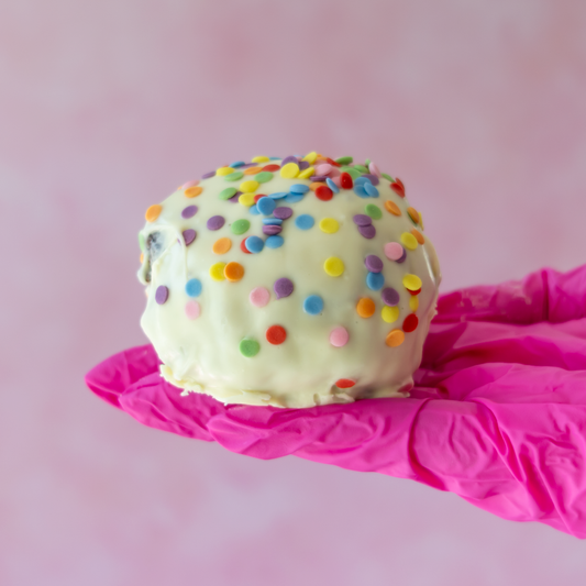 A spherical white chocolate-covered Brookie Choccy Boom Bite in colourful round sprinkles, held on a bright pink gloved hand against a soft pink background.