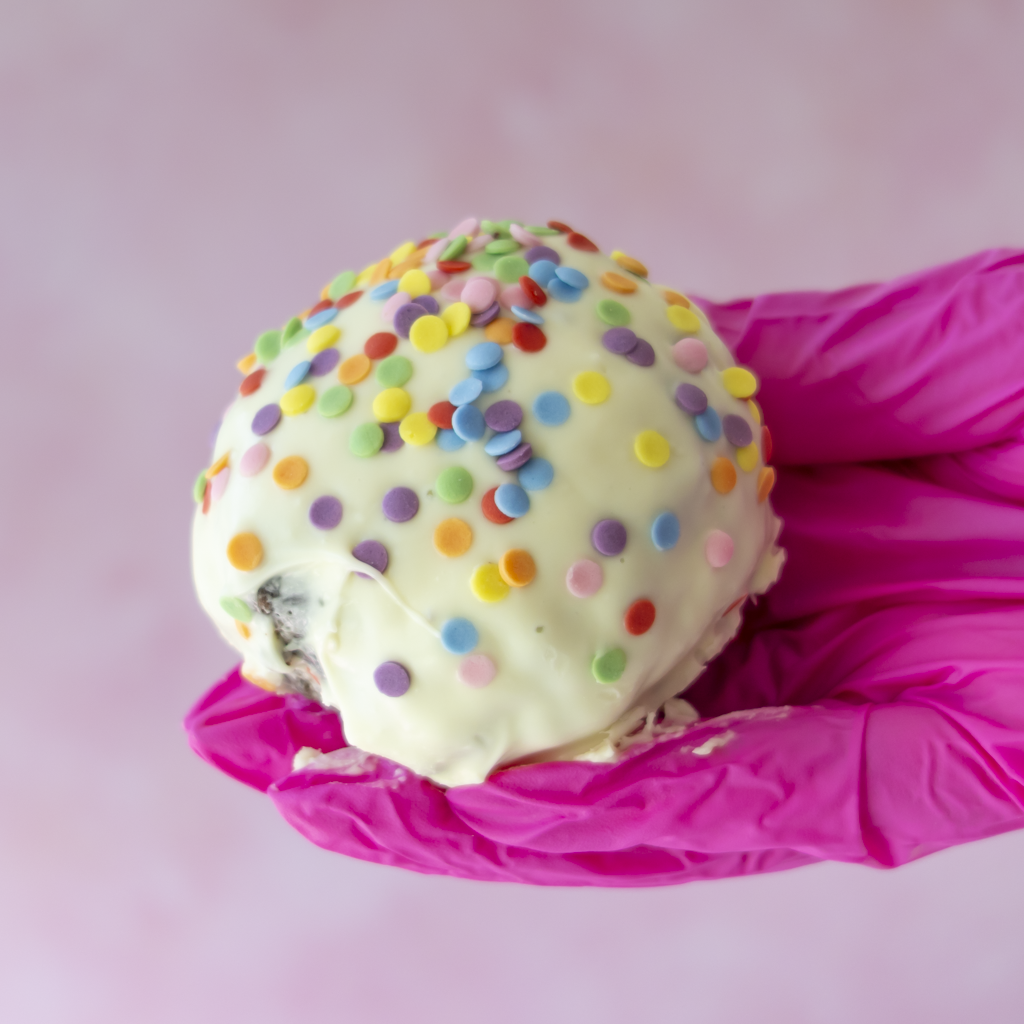 A white chocolate-covered Brookie Choccy Boom Bite topped with colourful round sprinkles, held in a bright pink gloved hand against a soft pink background.