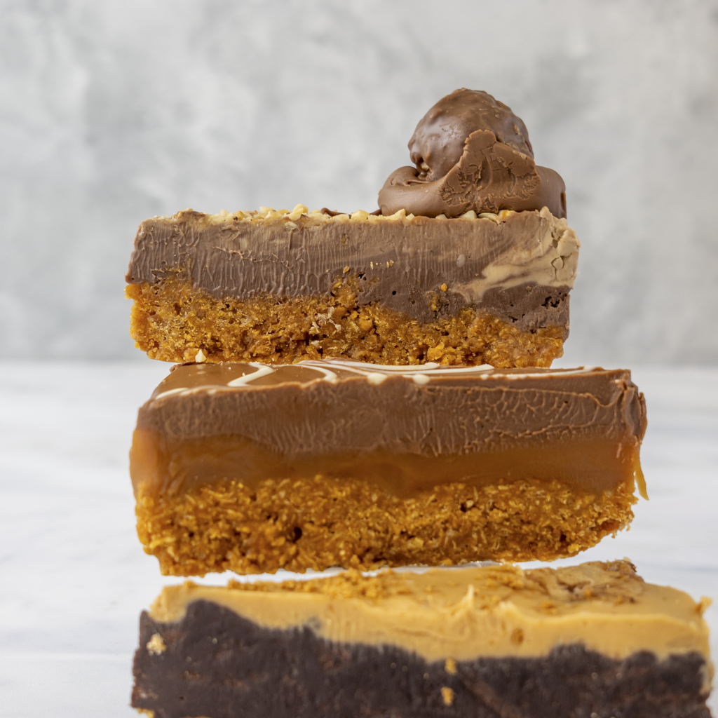 Three layered dessert bars by DoughGirl UK stacked on a light gray background