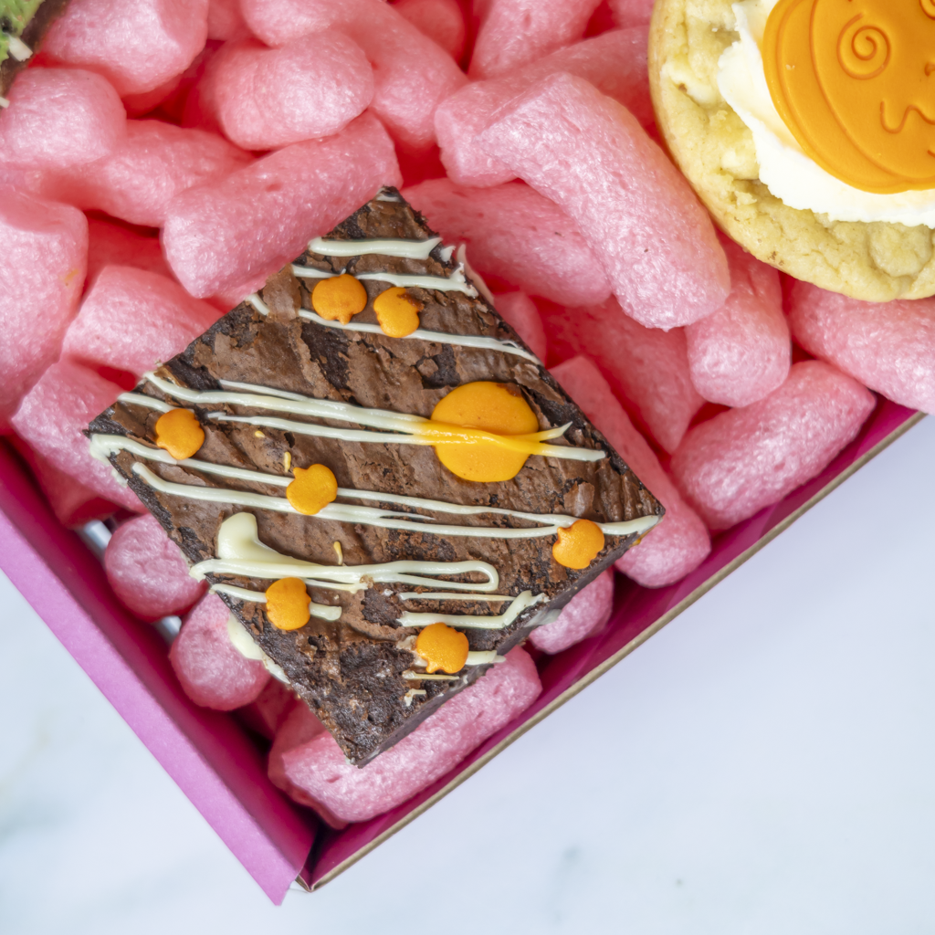 A close-up overhead shot of a chocolate brownie decorated with white drizzle and orange dots, presented in a pink gift box with pink packing peanuts and a cookie.