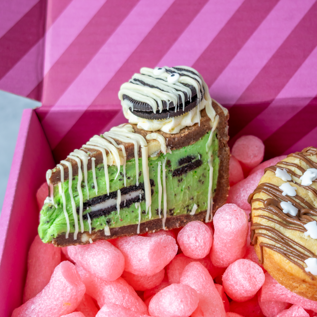 A slice of Midnight Monster Cookie Pie Slice with Oreo cookie layers and white chocolate drizzle, presented in a DoughGirl pink gift box.