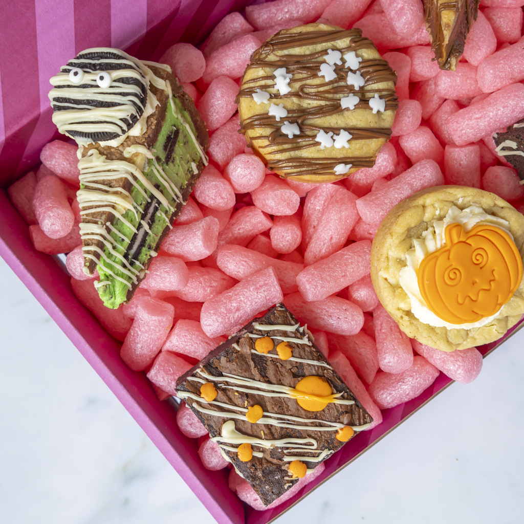 A boxed assortment of Halloween-themed baked goods, including a mummy cookie, a pumpkin cookie, and chocolate-dipped treats, arranged in pink packing foam inside a pink box.
