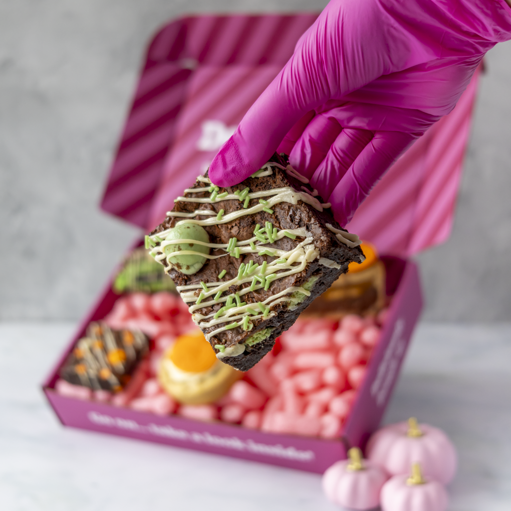 A hand wearing a bright pink glove holds a decorated brownie above an open pink gift box filled with cookies, slabs, and cookie pie slices, set on a light marble surface.