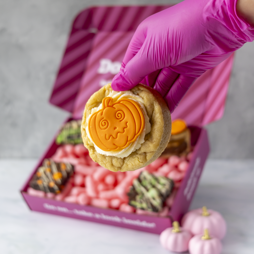 A hand wearing a bright pink glove holds a decorated pumpkin-shaped cookie in front of an open pink gift box filled with candy and treats. Small pink pumpkins sit on the surface nearby.