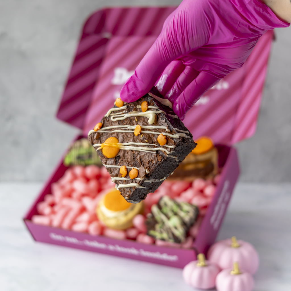 A hand wearing a bright pink glove holds a square brownie decorated with white drizzle, orange chocolate pieces, and chocolate chips above an open pink box filled with assorted treats.