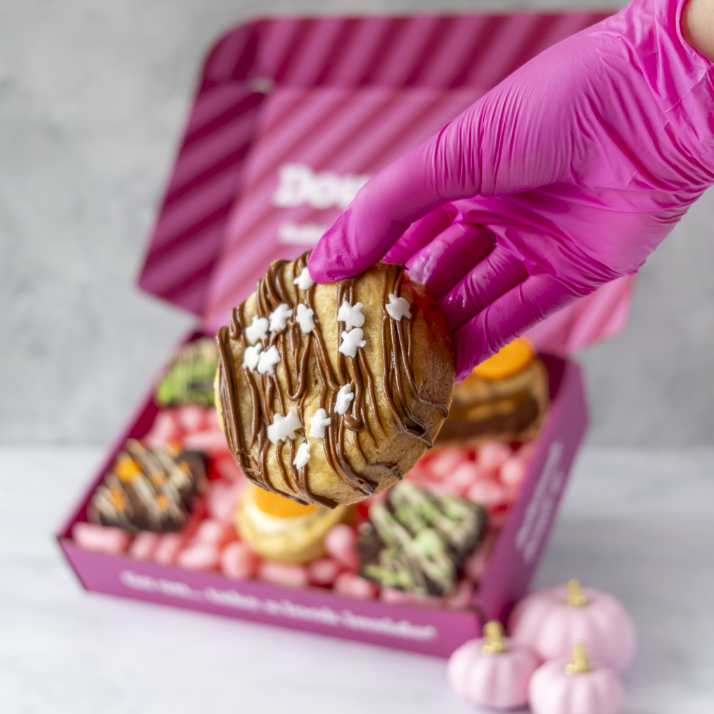 A hand wearing a bright pink glove holds a decorated chocolate chip cookie in front of an open pink gift box filled with assorted cookies on a light gray surface.