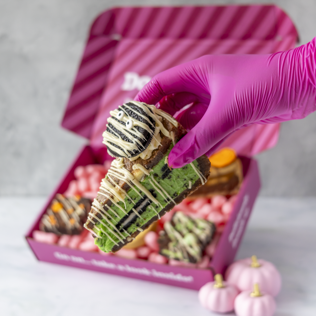 A hand wearing a bright pink glove holds a decorated chocolate cookie pie piece in front of an open pink gift box and assorted treats. Small pink pumpkins sit on the surface in the foreground.