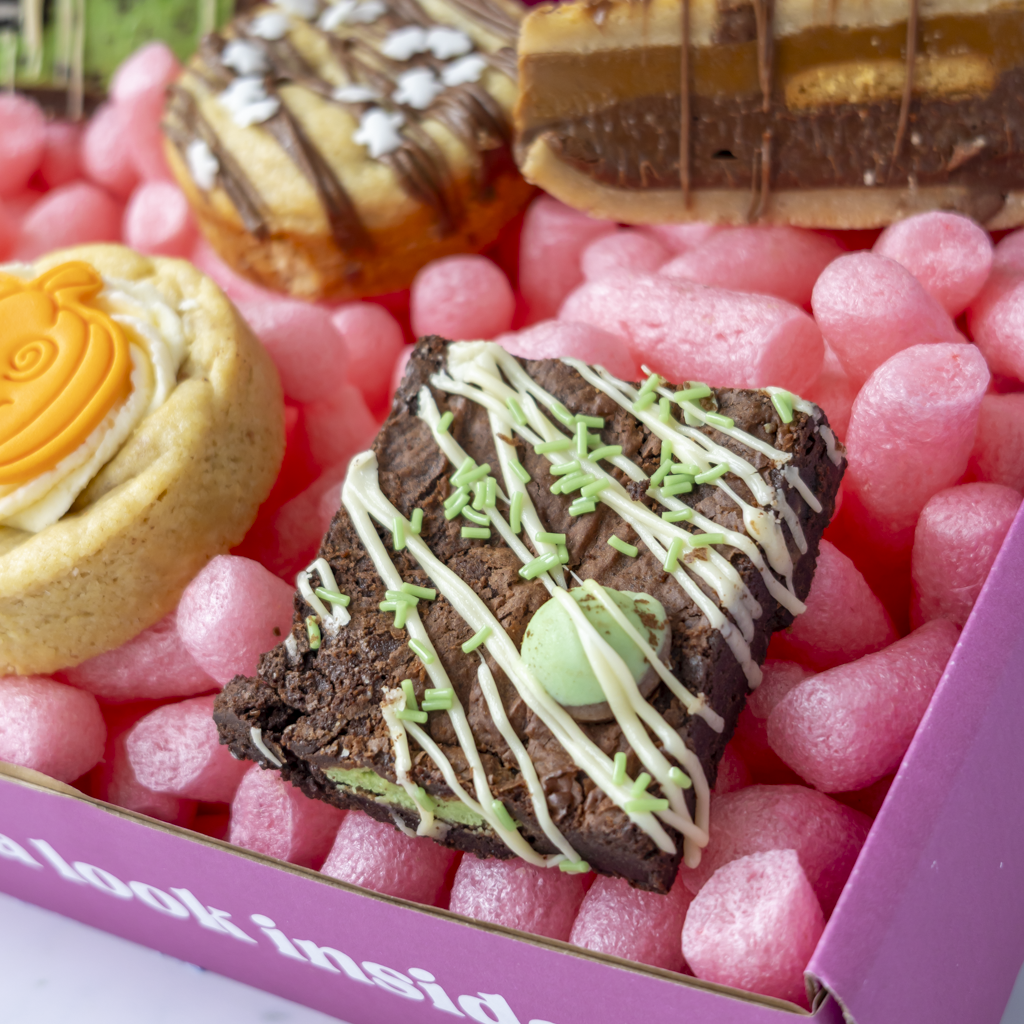 A close-up of assorted baked goods arranged in a pink packing bubble box. The most prominent item is a square chocolate brownie topped with white drizzle, green sprinkles, and a mint candy.