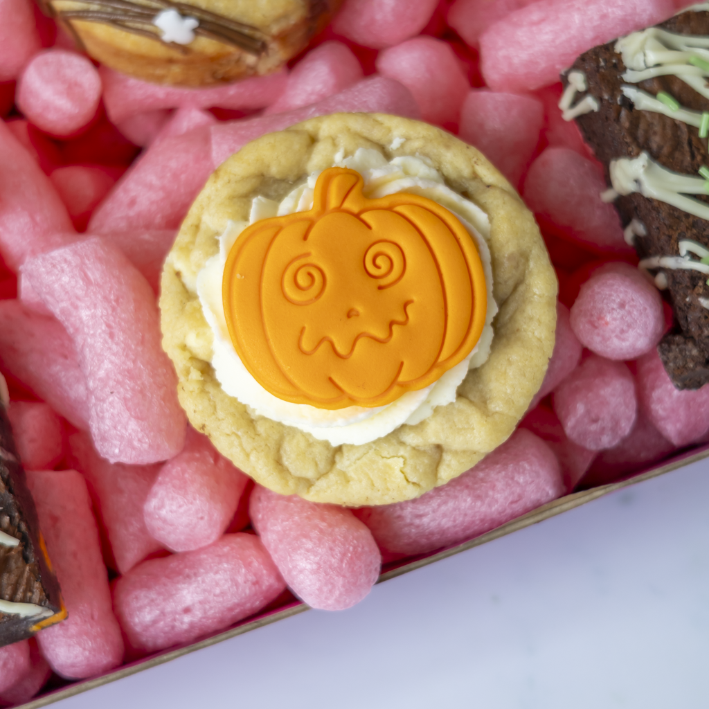 A close-up of a round baked cookie topped with white frosting and an orange pumpkin-shaped decoration, resting on pink packing peanuts inside a box.