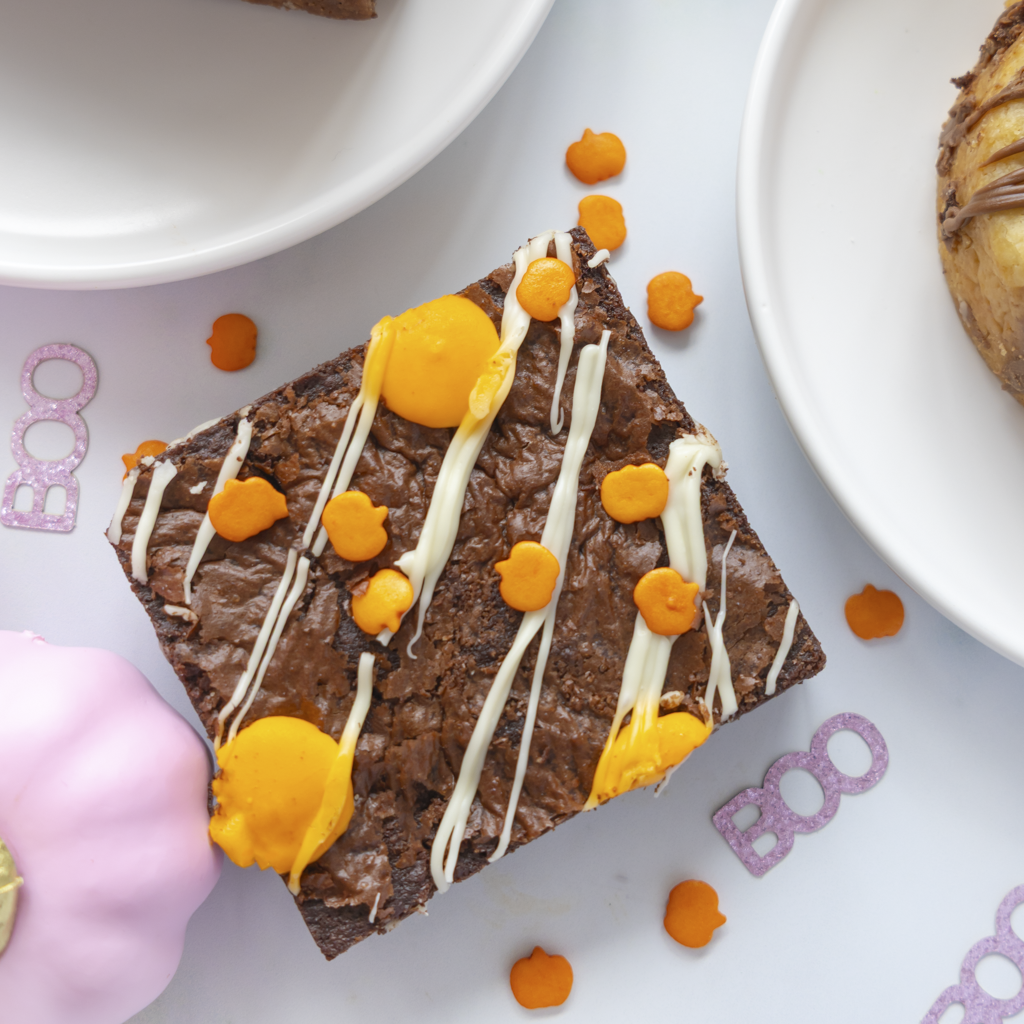 A square chocolate brownie topped with white chocolate drizzle, bright orange sweet pieces, and two large orange sweet drops, photographed on a white surface with partial plates and decorative pink accents around it.