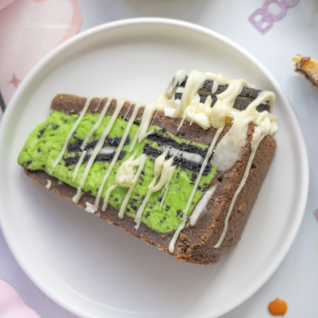 A slice of layered cookie pie on a white plate, featuring a dark chocolate base, a bright green middle layer, and a top layer with white drizzle and dark cookie strips.