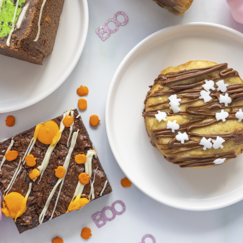 Treat-themed Halloween desserts, including chocolate brownies with orange and white drizzle and a glazed cookie on white plates, with 'BOO' decorations on a light background.