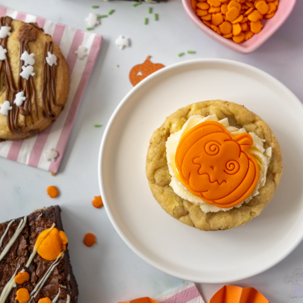 A Halloween-themed cookie topped with white frosting and an orange pumpkin-shaped decoration, served on a white plate. Other decorated cookies and orange sprinkles are visible in the background on a light surface.