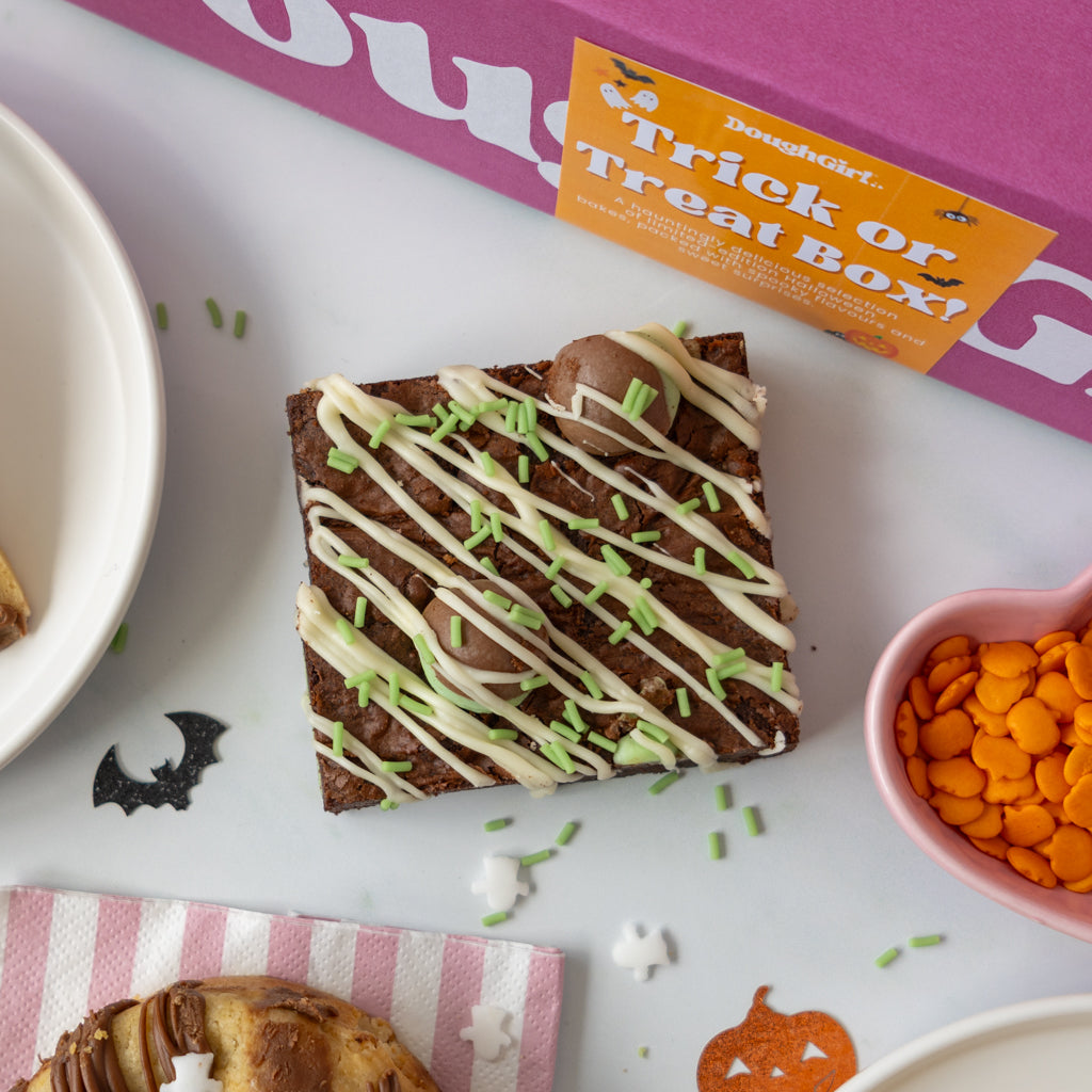 A Halloween-themed treat box setup on a white surface, featuring a chocolate brownie with white and green drizzle, alongside an orange 'Treat or Trick Box!' card, a pink bowl of orange candy corn, and partial plates and pastry.