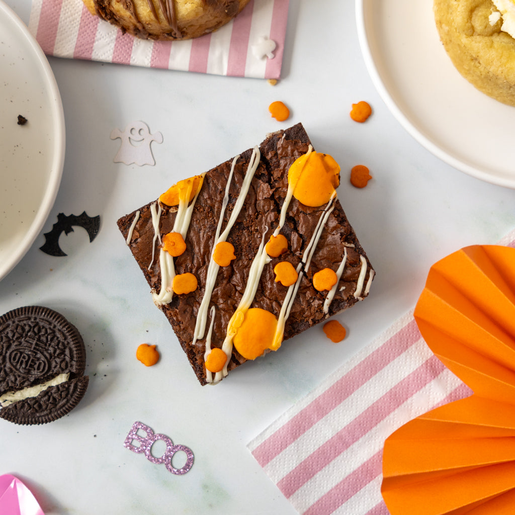 A Halloween-themed chocolate brownie decorated with white drizzle, orange candy corn, and orange sweet dots, styled on a light marble surface with Oreo cookies, cookies and cream, and orange paper decorations.