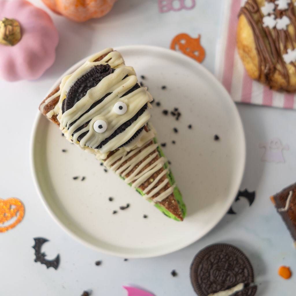 A Halloween-themed slice of cake on a white plate, decorated with white icing, chocolate drizzle, Oreo cookies, and sweet eyes, surrounded by autumn decorations.