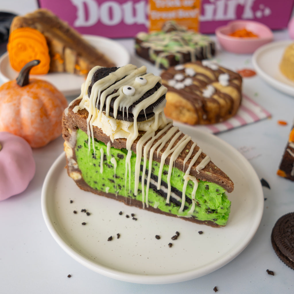 A Halloween-themed slice of cookie pie on a white plate, decorated with green frosting, white drizzle, and an Oreo cookie on top, surrounded by pumpkins and other decorated bakes.