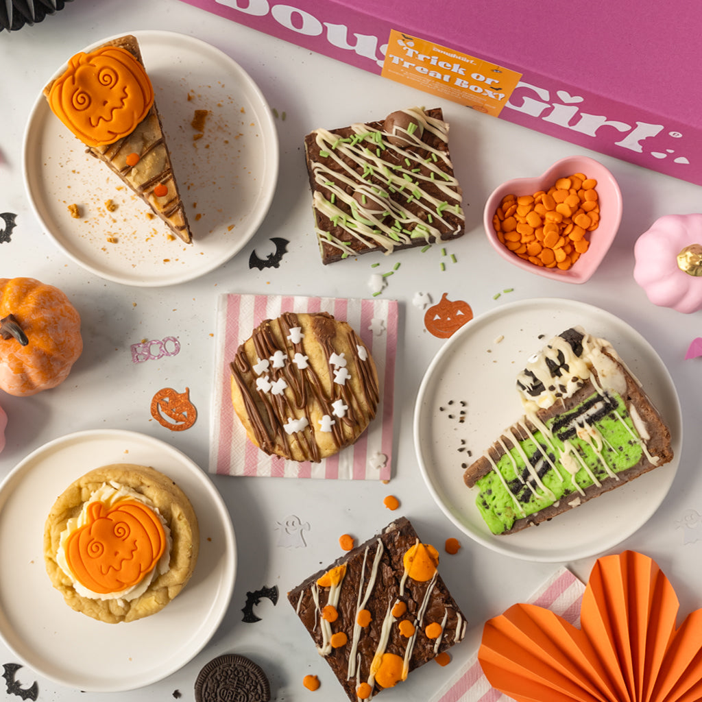 A styled flat lay of Halloween-themed dessert treats on white plates, including pumpkin cookie pie slices, chocolate brownies with drizzles, and cookies, arranged with orange paper fans, a pink heart dish, and a pink box labelled 'DoughGirl' on a white surface.