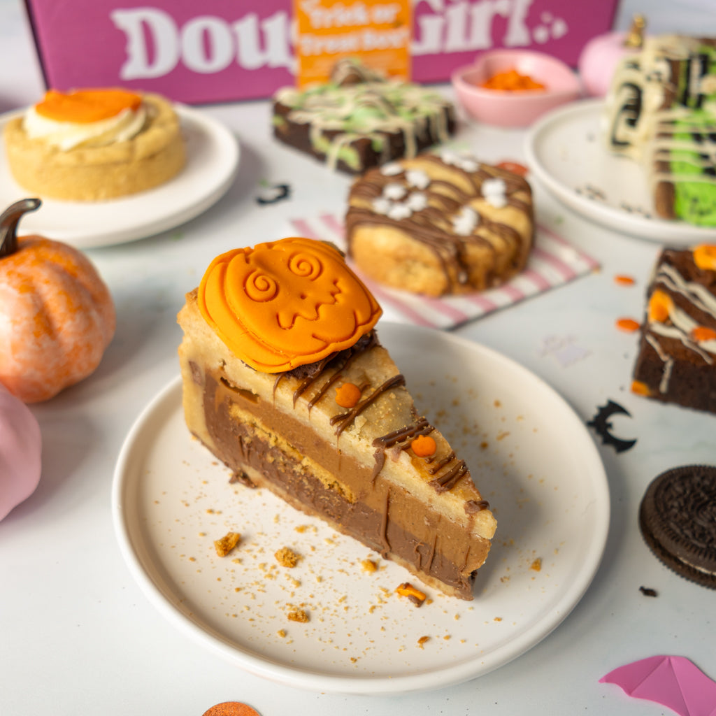 A slice of layered pumpkin cookie pie with a carved orange pumpkin topper sits on a white plate, surrounded by Halloween-themed desserts, mini pumpkins, and candy on a decorated table.