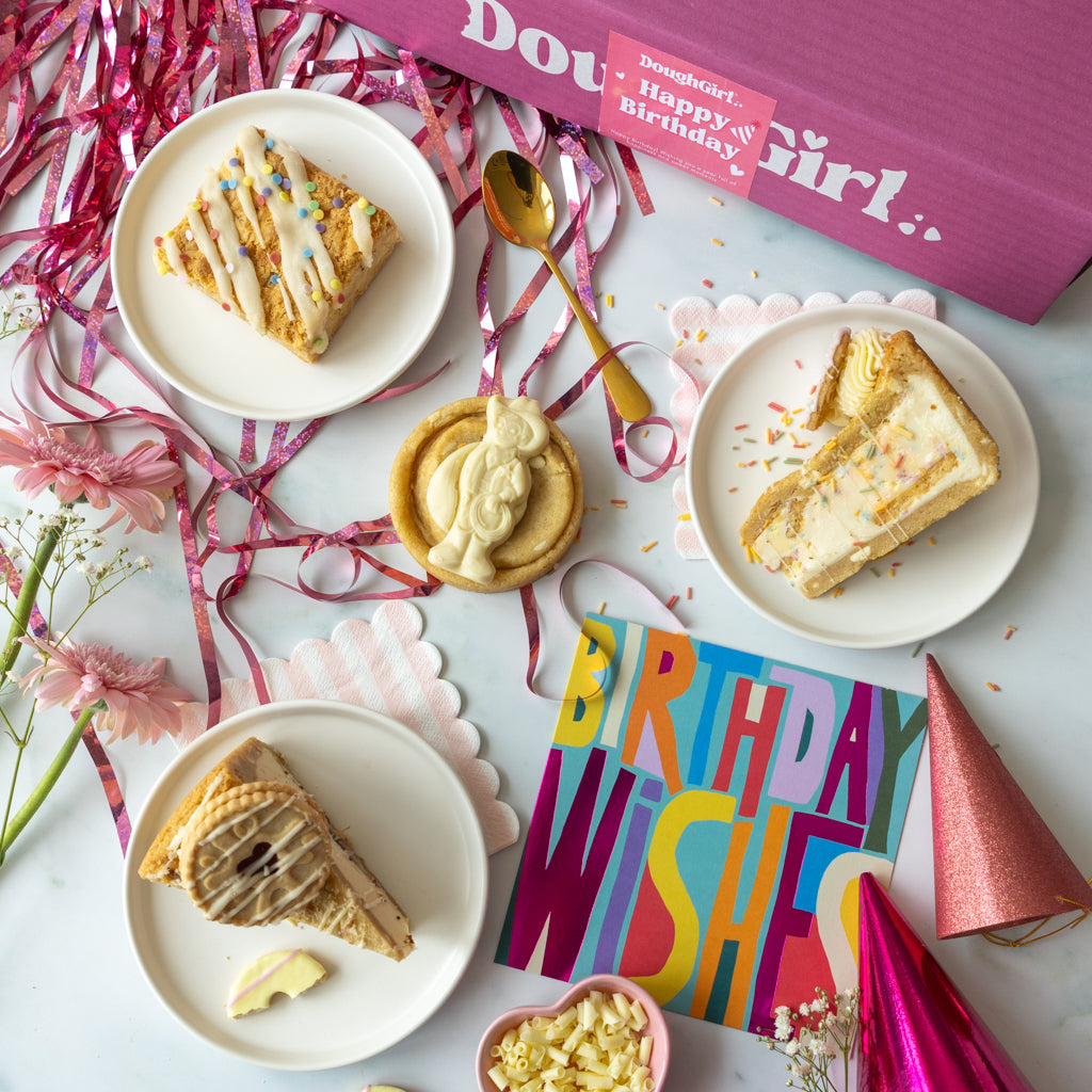 A styled birthday gift scene with three slices of cookie pie slices and a slab on white plates, a small cookie, a colorful 'Birthday Wishes' card, pink party hats, confetti, streamers, and a pink box labeled 'DoughGirl, Happy Birthday!'.
