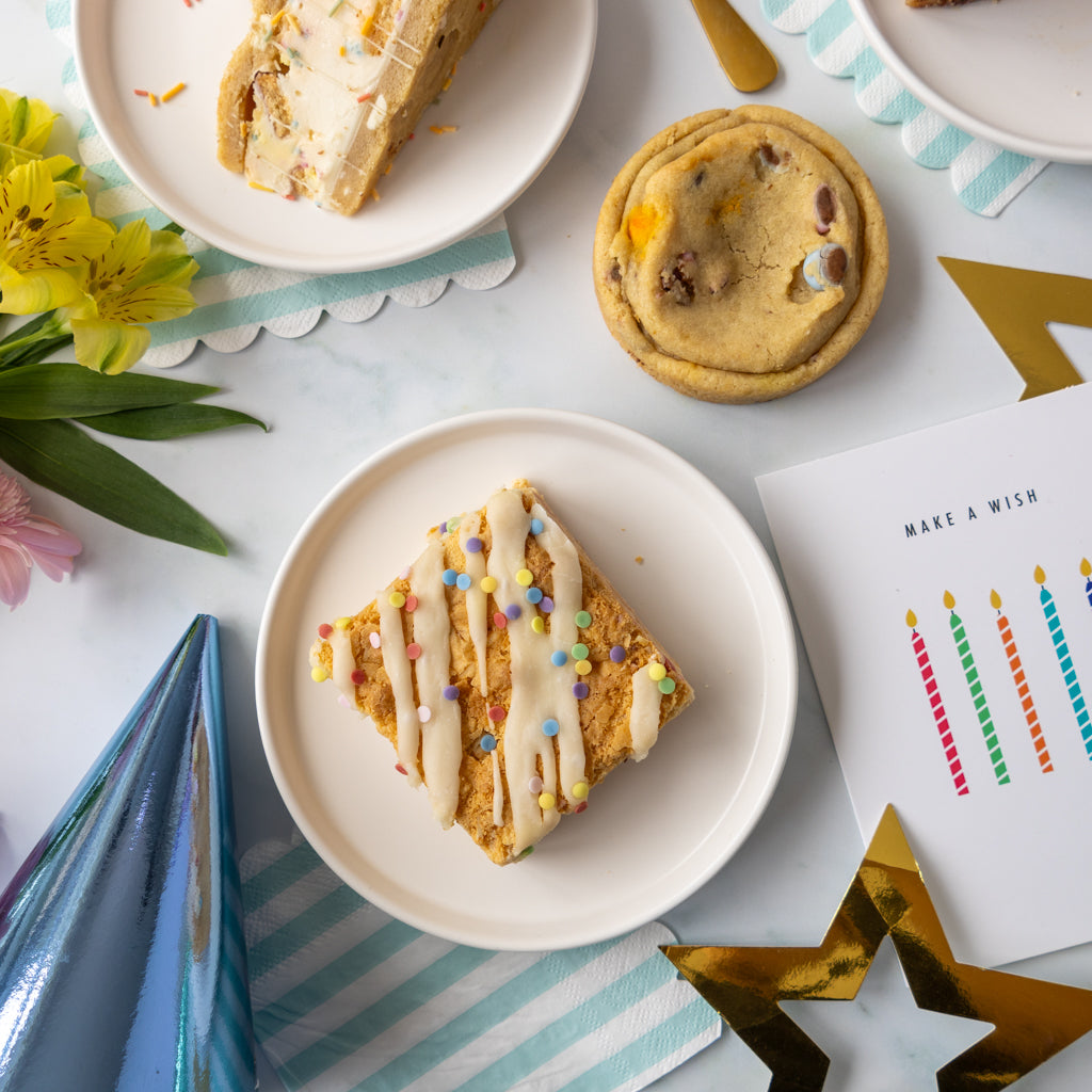 A styled flat-lay of birthday treats on a white surface, featuring a square blondie slab with white icing and colorful sprinkles on a plate, a chocolate chip cookie, a slice of layered cake, party hats, a star decoration, flowers, and a card that says 'MAKE A WISH'.