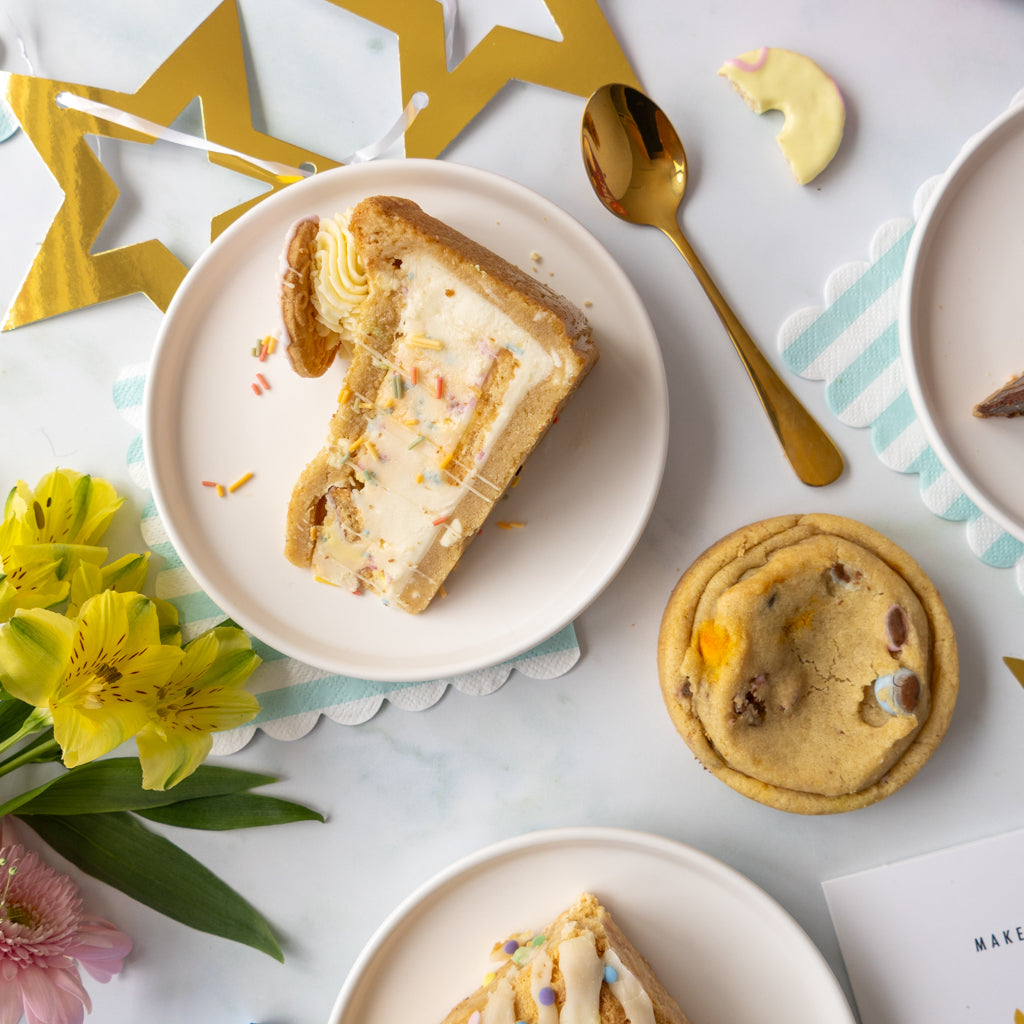 A styled flat-lay of baked bakes on a white marble surface, featuring a slice of layered cookie pie on a white plate, a chocolate bean cookie, a gold spoon, yellow flowers, and gold star decorations.