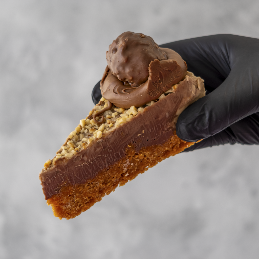 Slice of chocolate cake with hazelnut being held by a gloved hand against a neutral background