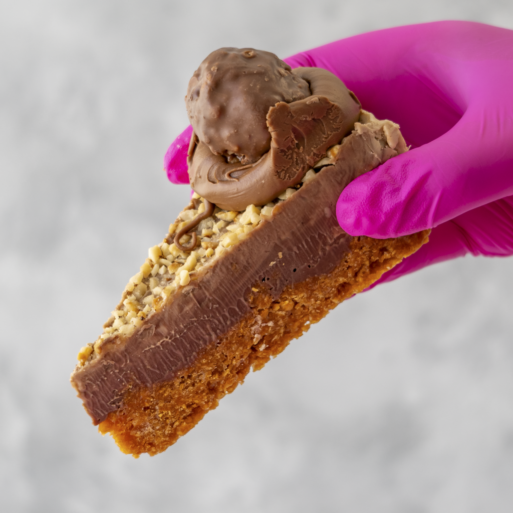 DoughGirl Hazelnut Cornflake Pie Slice with a hazelnut truffle held by a pink gloved hand against a gray background