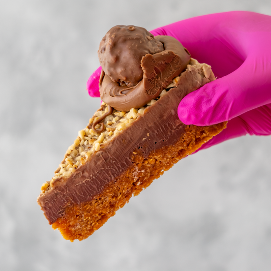 DoughGirl Hazelnut Cornflake Pie Slice with a hazelnut truffle held by a pink gloved hand against a gray background