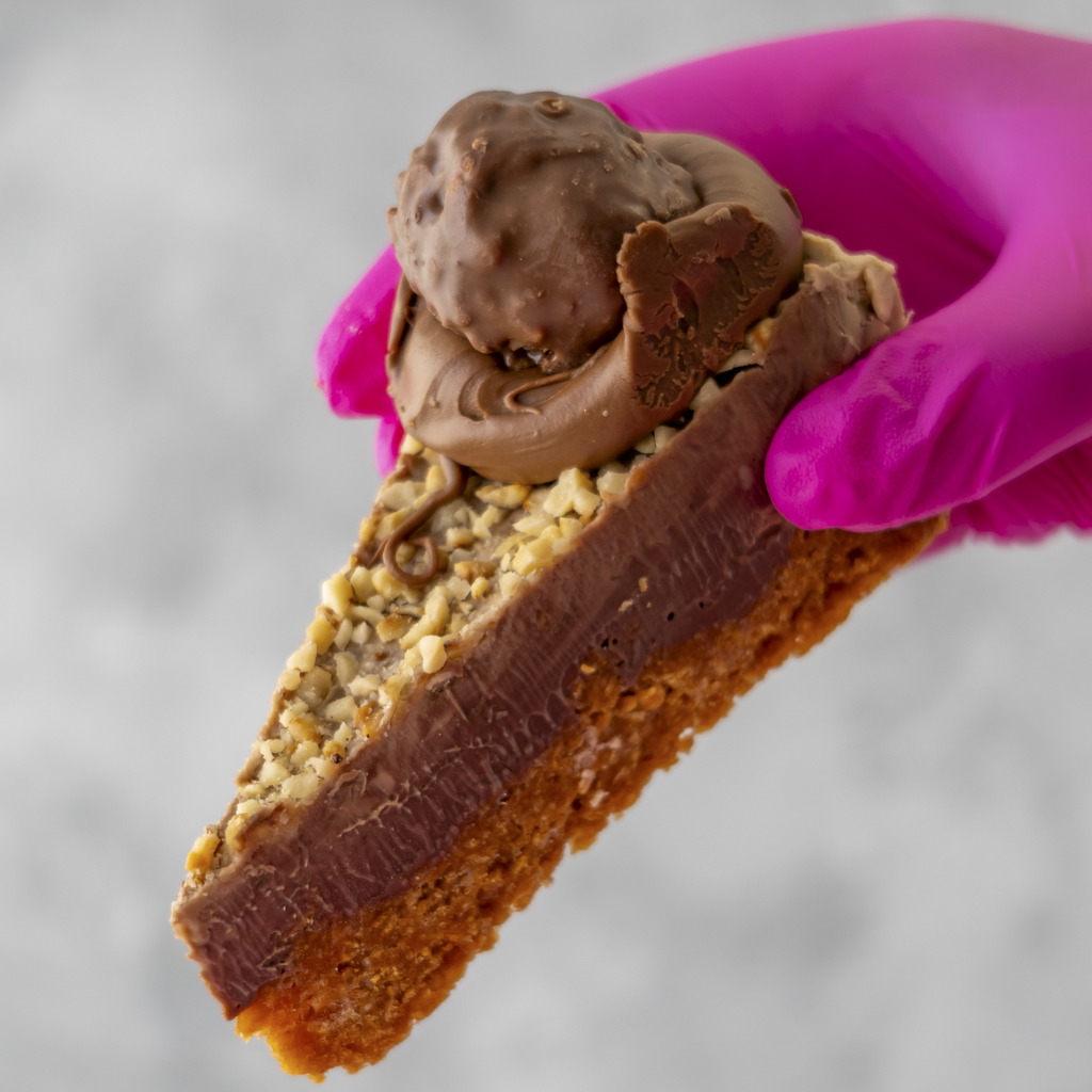Hazelnut Cornflake Pie Slice by DoughGirl UK held by a hand wearing a pink glove against a neutral background