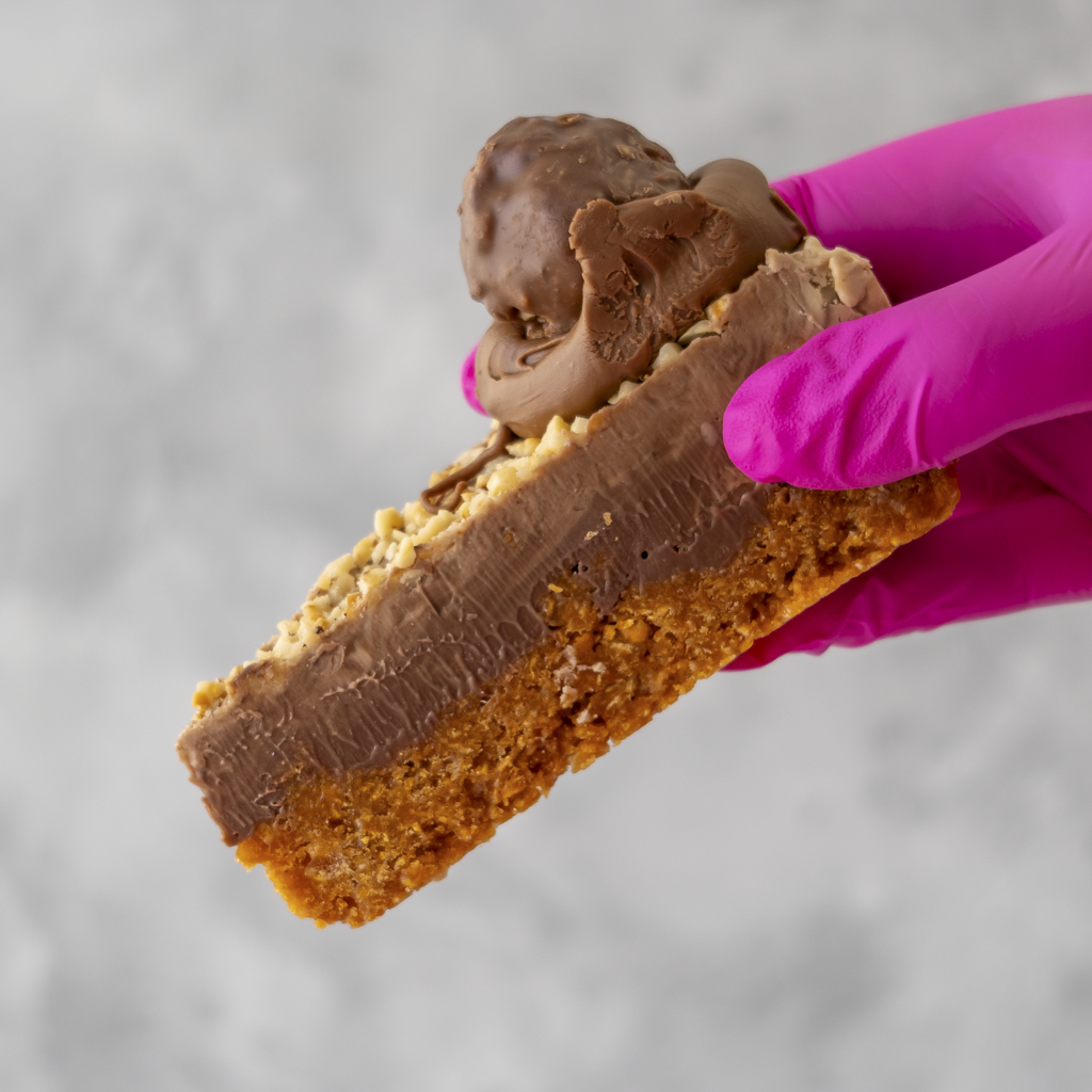 Hazelnut Cornflake Pie Slice with a cornflake base held by a hand wearing a pink glove on a gray background