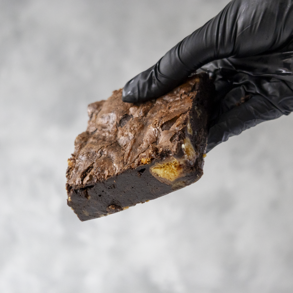 Hand wearing a black glove holding a Honeycomb Crunch Brownie Slab against a gray background