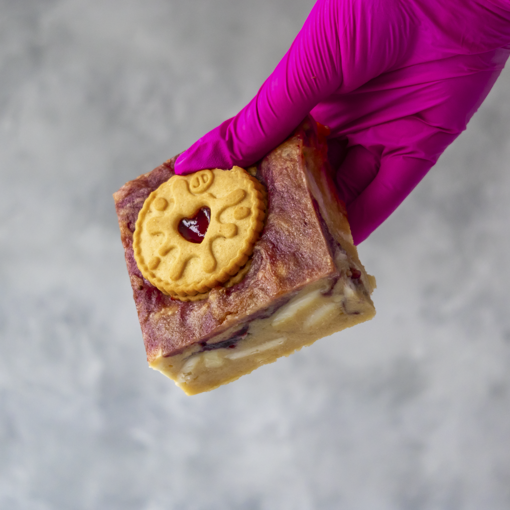 Hand wearing a pink glove holding a Jammie Dodger Blondie Slab with a Jammie Dodger cookie on top against a gray background
