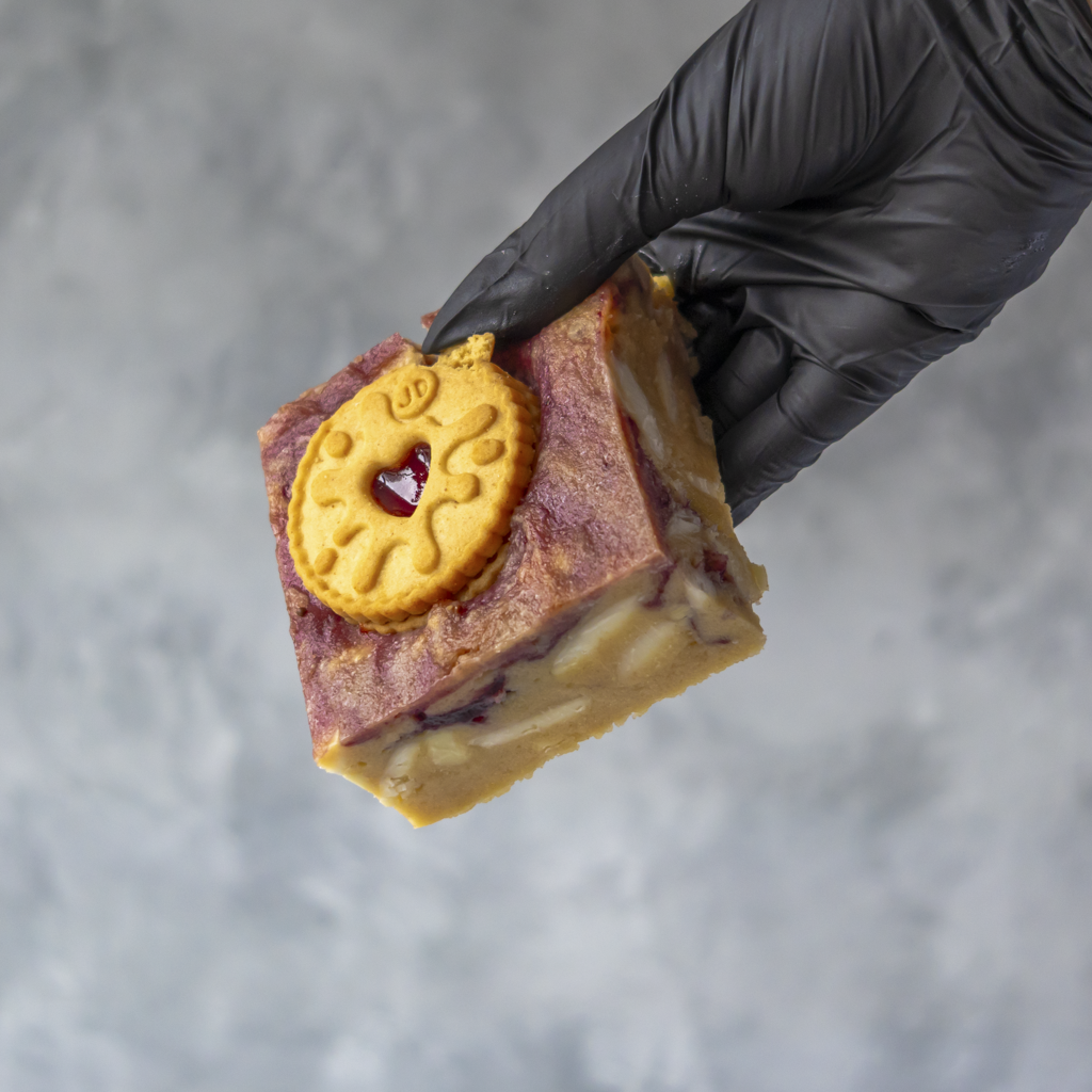 Hand holding a DoughGirl Jammie Dodger Blondie Slab with a Jammie Dodger biscuit on top against a gray background