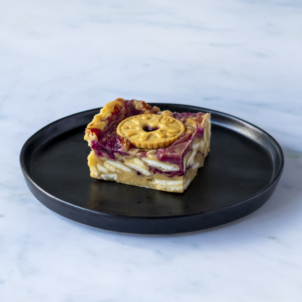 Jammie Dodger Blondie Slab by DoughGirl UK with a Jammie Dodger biscuit on top on a black plate against a light background