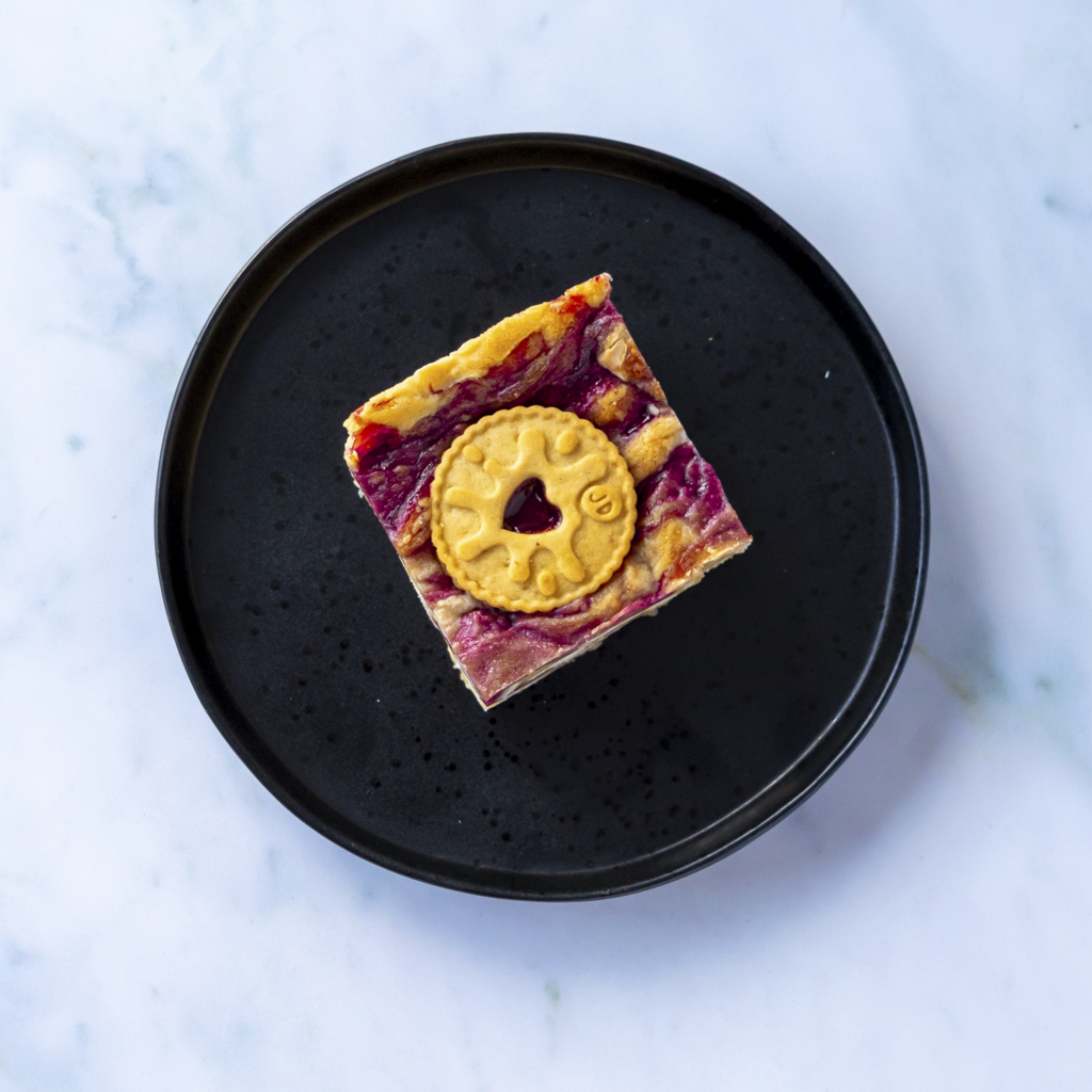 Square of Jammie Dodger Blondie Slab with round biscuit center on a black plate against a light blue marble background, perfect for dessert and sweet treat