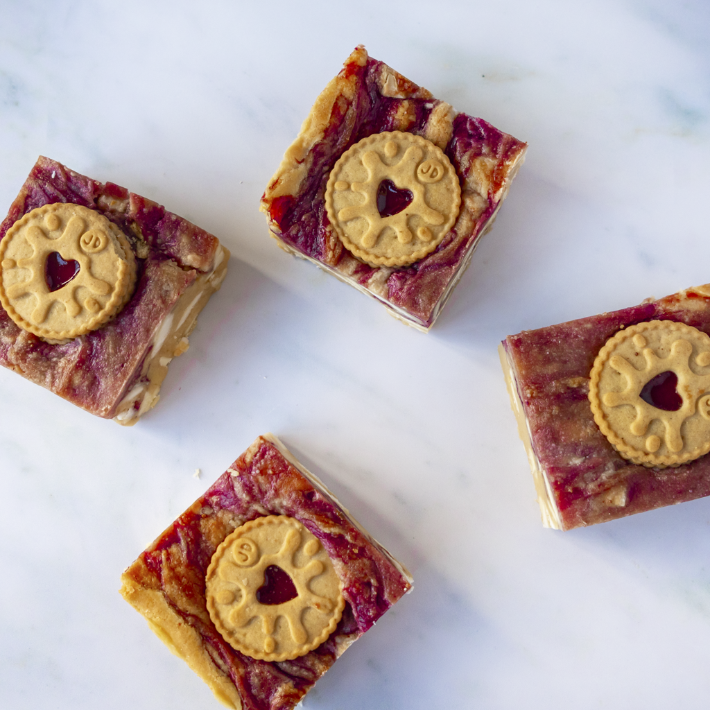 Square Jammie Dodger Blondie Slab with a red fruity jam filling and a classic Jammie Dodger biscuit on top, arranged on a white marble surface.