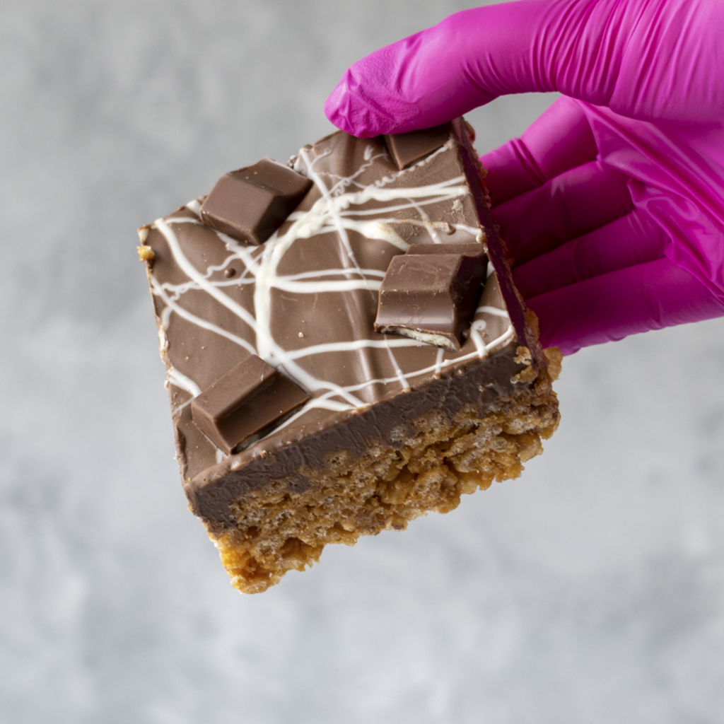 Hand holding a piece of chocolate dessert Kinder Crispie Slab with a purple glove on a gray background
