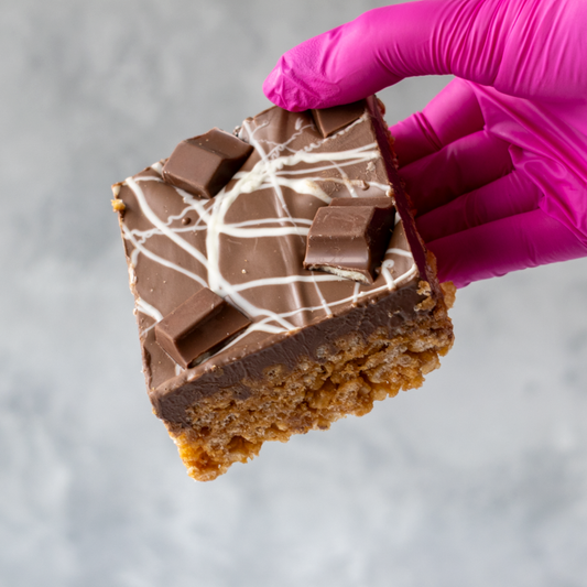 Hand holding a piece of chocolate dessert Kinder Crispie Slab with a purple glove on a gray background