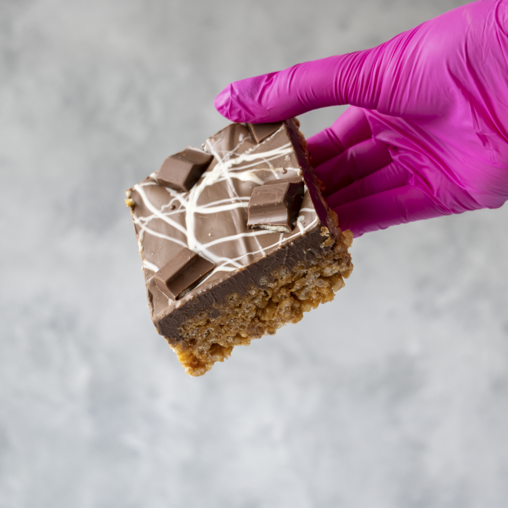 Hand wearing a purple glove holding a piece of Kinder Crispie Slab against a gray background