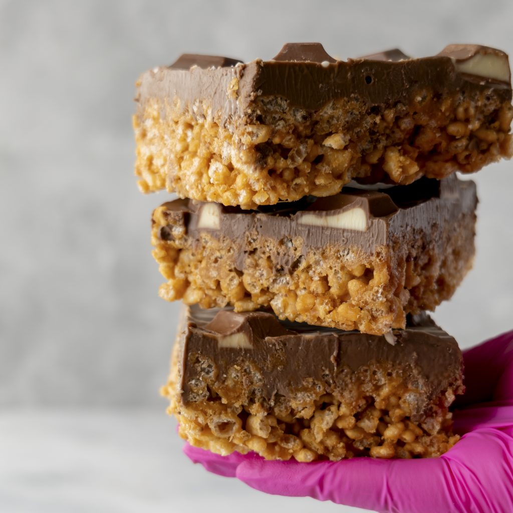 Stacked Kinder Crispie Slab slices held in pink glove showing caramel crispie base, chocolate layer and Kinder chunks