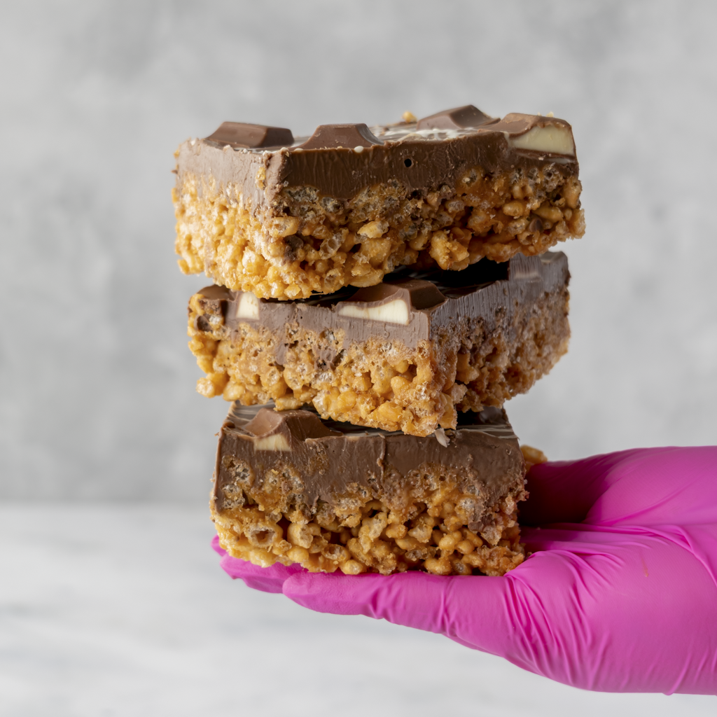 Three layers of Kinder Crispie Slab dessert with Kinder chocolate pieces and white chocolate drizzle held in gloved hand