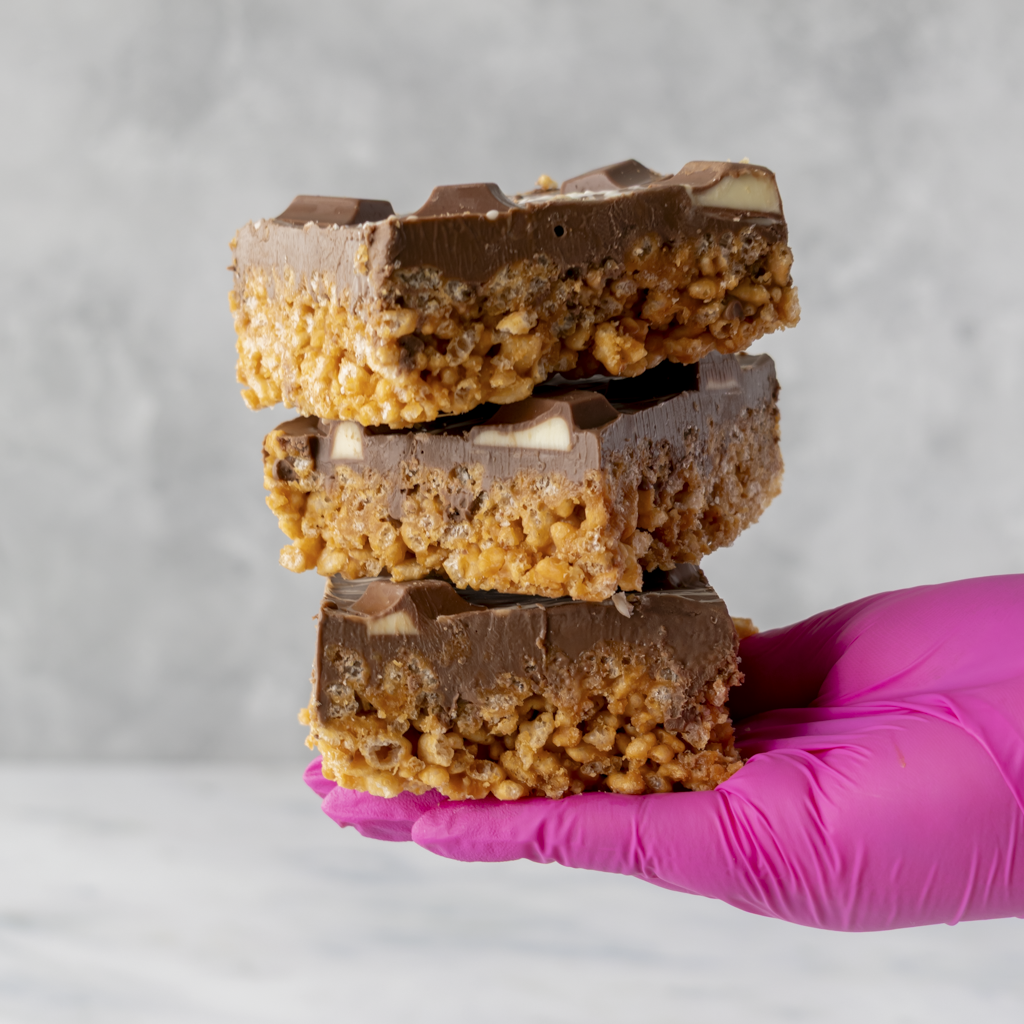 Hand holding three layered Kinder Crispie Slabs with caramel rice crispy base, hazelnut chocolate and Kinder bar pieces on grey background