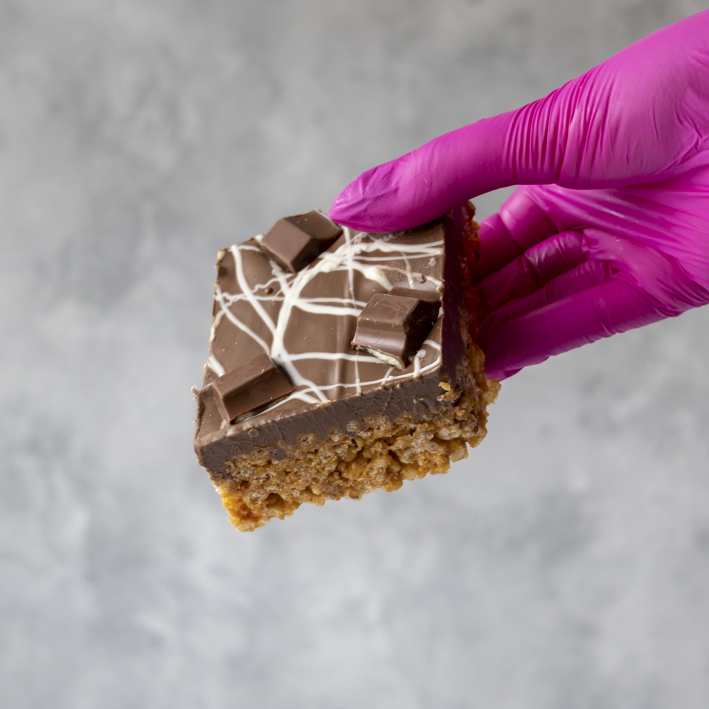 Chocolate-covered Kinder Crispie Slab held by a hand wearing a purple glove on a gray background