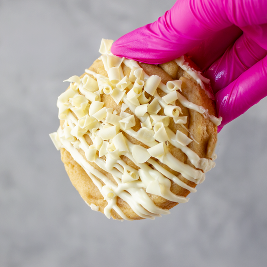 Lemon & White Chocolate Stuffed Deluxe Cookie with white chocolate drizzle held by a hand wearing a pink glove on a gray background