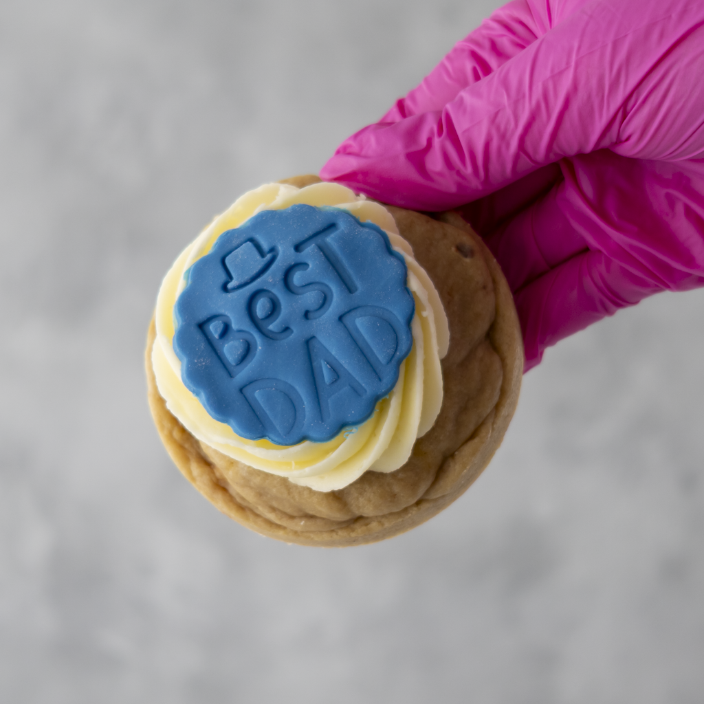Father's day cookie with 'Best Dad' stamp held by a gloved hand against a gray background