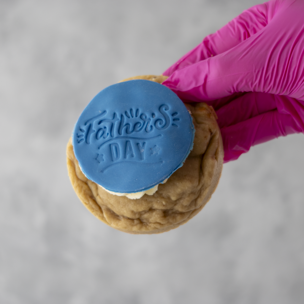 DoughGirl Cookie with blue 'Father's Day' decoration held by a pink-gloved hand on a gray background