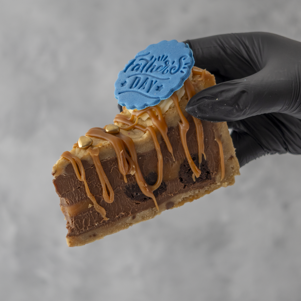 Slice of Father's Day Salted Caramel Brownie Cookie Pie with caramel drizzle and 'Father's Day' topper held by a gloved hand against a gray background