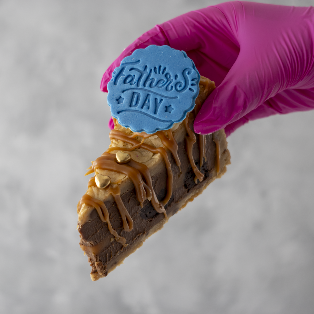 Slice of cookie pie with 'Father's Day' stamp held by a gloved hand on a gray background