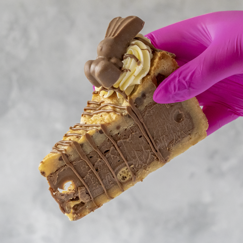 Limited Edition Malteser Bunny Cookie Pie Slice with chocolate drizzle and a chocolate bunny held by a hand wearing a pink glove on a light gray background.